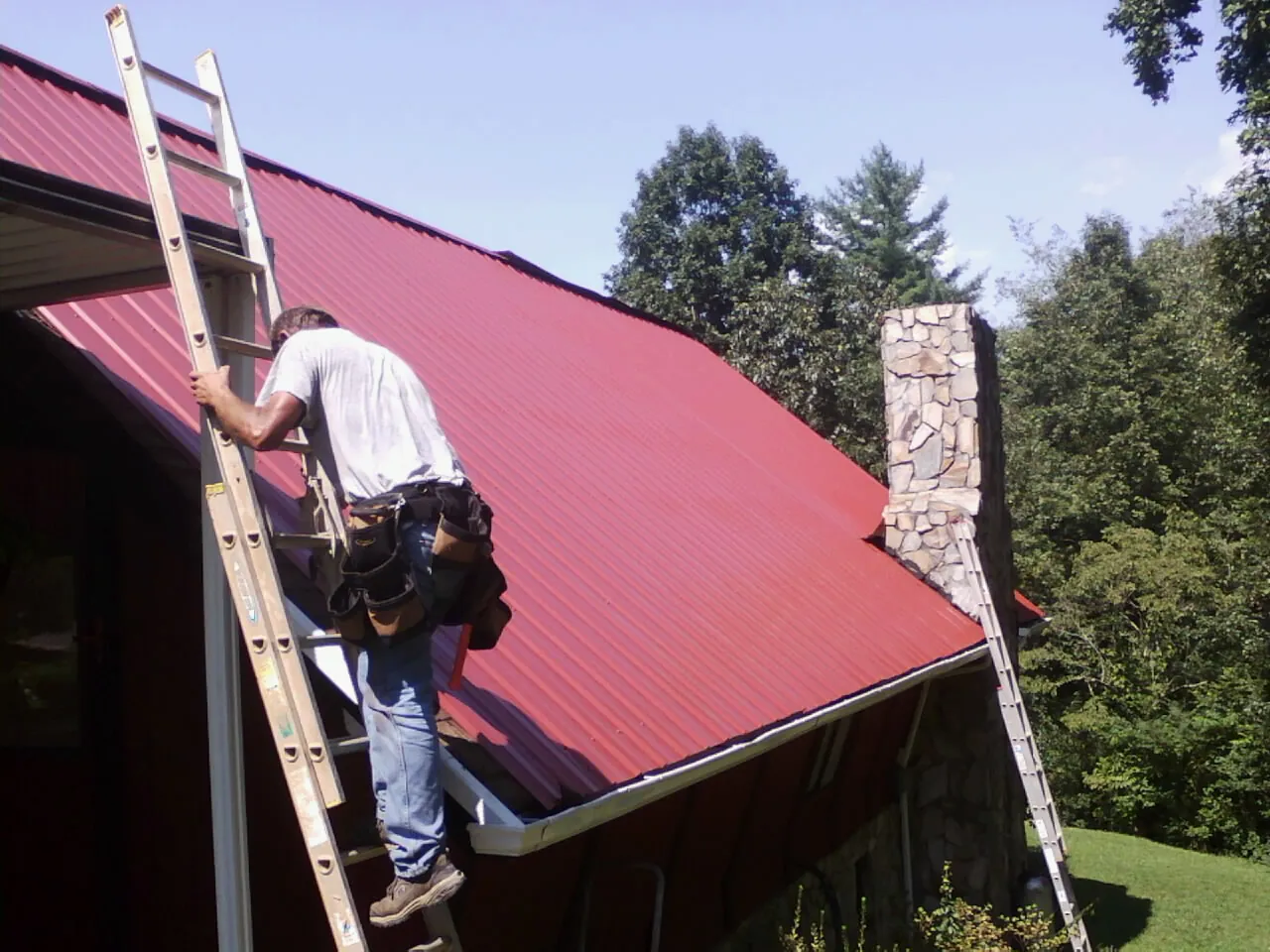 Roofer on a ladder beside a red metal roof and stone chimney — Soffit & Fascia Repair by Roofing Brothers in Media